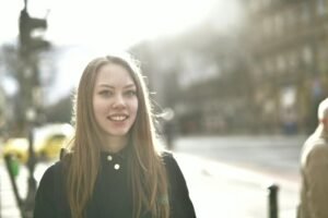 A young woman smiling brightly outdoors on a sunny street.